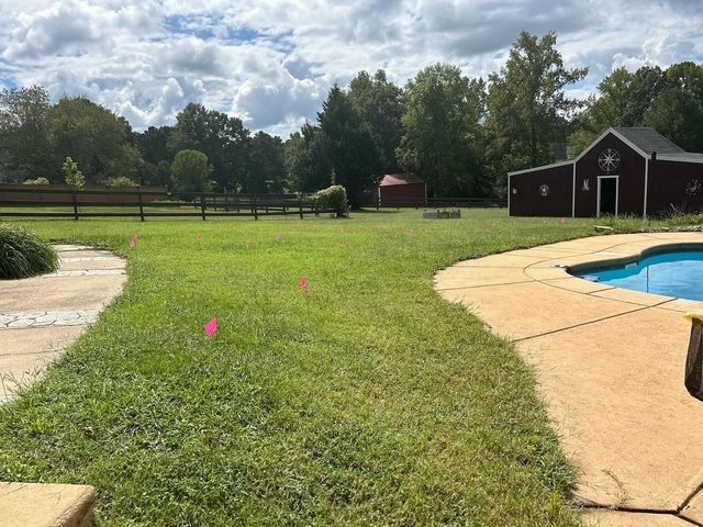 Backyard lawn with pool, patio, sheds, and pink markers under cloudy sky