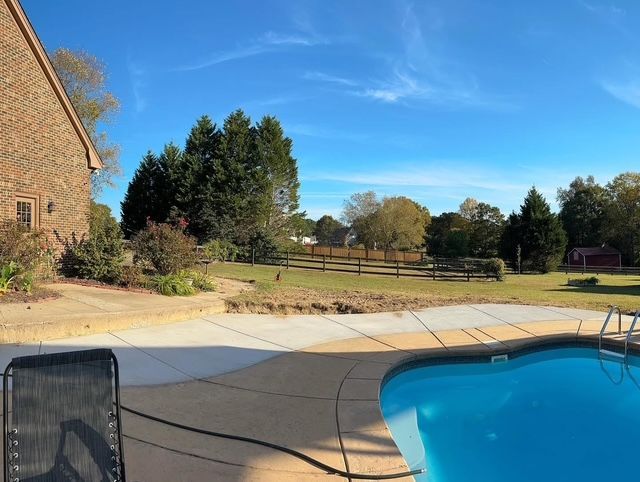 Backyard patio with a swimming pool, lawn, trees, and a brick house under a clear blue sky