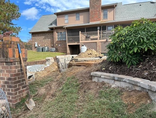 Backyard excavation beside a brick house, with exposed dirt, retaining walls, and construction debris