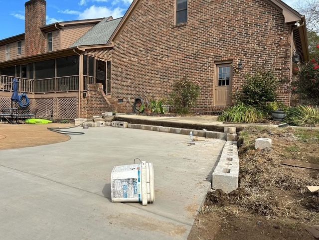 Brick house with a concrete driveway, retaining wall blocks, and a small bucket in the foreground.