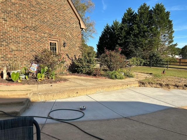 Backyard patio beside a brick house, with garden beds, shrubs, and a lawn under clear blue sky.