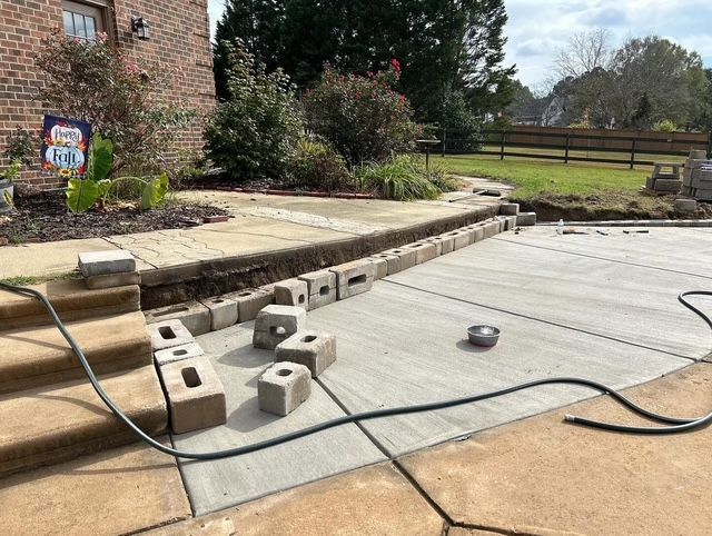 Construction site with concrete patio, cinder blocks, and hose beside a brick house and landscaped yard