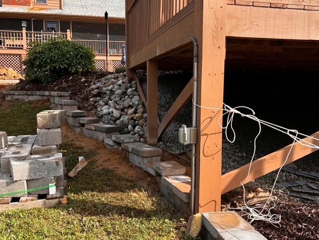 Damage under wooden deck with collapsed stone retaining wall and scattered cinder blocks in a yard.