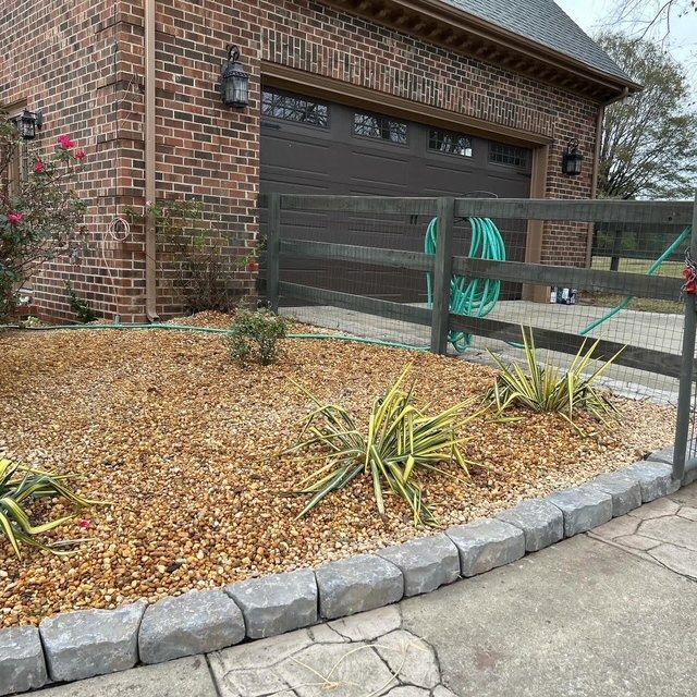 Front yard with brick house, garage door, curved stone border, and small landscaped bed with plants and gravel.