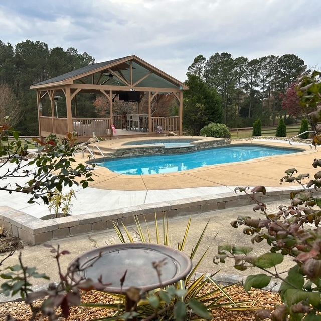 Backyard pool with stone deck, covered patio, and landscaped garden under cloudy sky