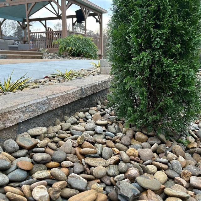 Landscaped yard with a small evergreen tree, river rocks, and a stone border by a patio and gazebo