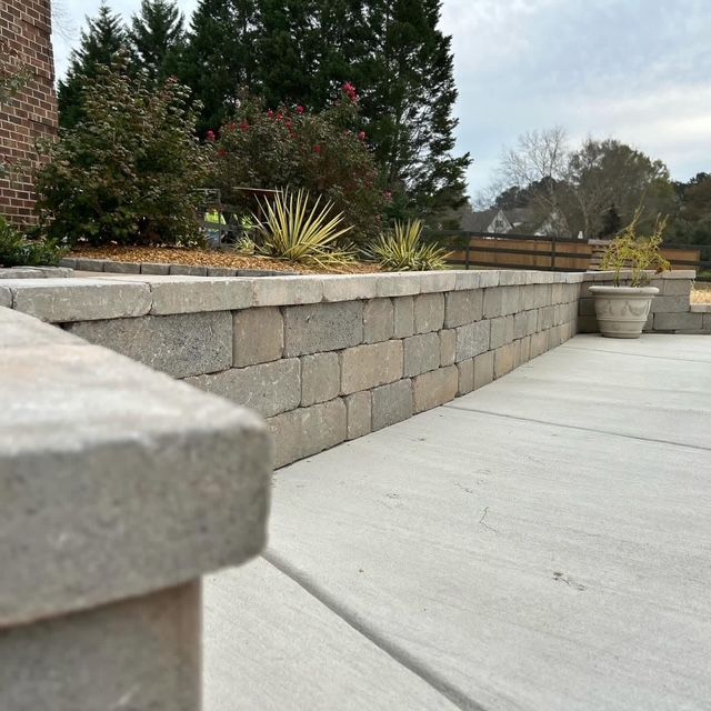 Stone retaining wall beside a concrete patio with potted plants and shrubs in a garden.