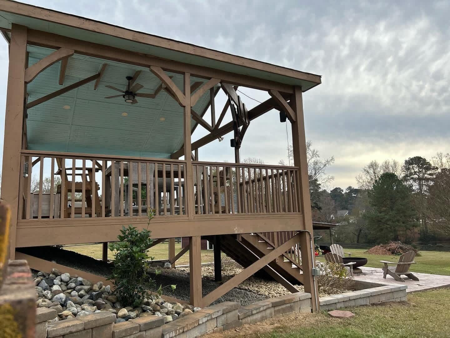 Wooden gazebo with railings and stone base in a landscaped yard under cloudy skies