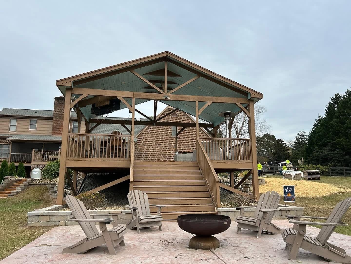 Rustic wooden gazebo with stairs, surrounded by Adirondack chairs and a fire pit outdoors.