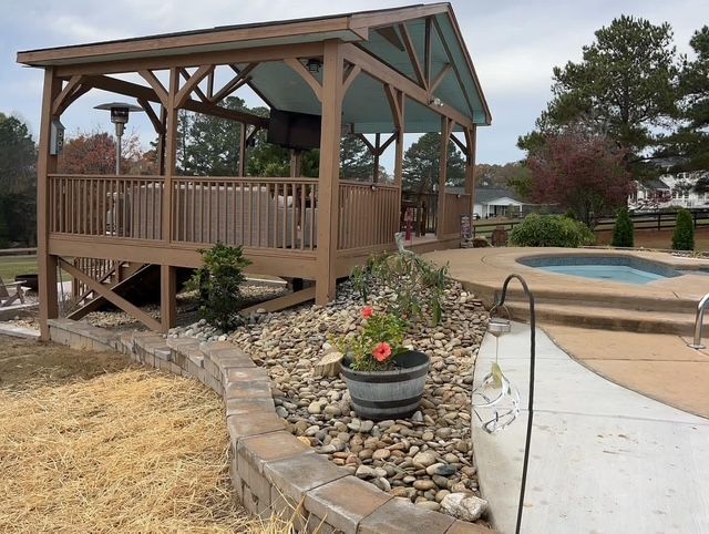 Wooden gazebo beside a landscaped pool area with rocks, flowers, and a concrete path.