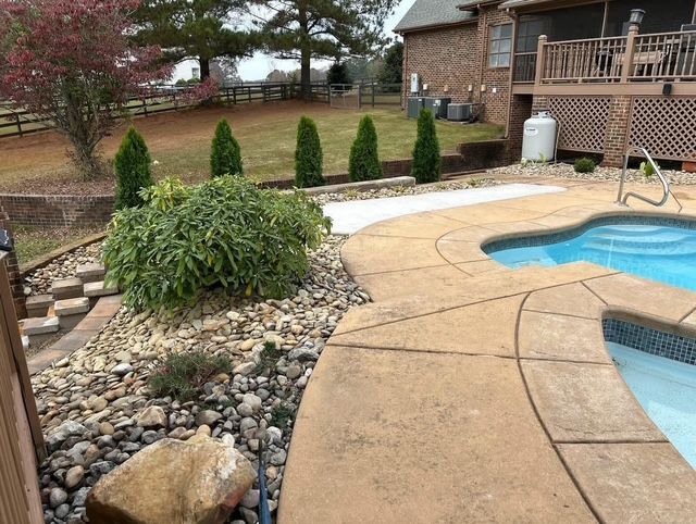 Backyard swimming pool with curved tan patio, landscaped rocks, shrubs, and a brick house in the background