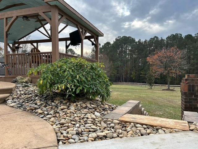 Patio and pavilion with a green shrub, stone landscaping, and grassy yard with trees beyond