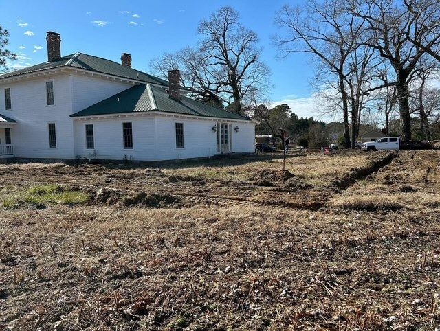 White house with a green roof in a bare field under a clear blue sky, with trees and a parked truck.