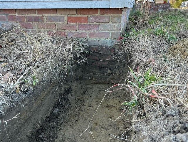Narrow muddy trench beside a brick house foundation with dry grass and exposed soil sides