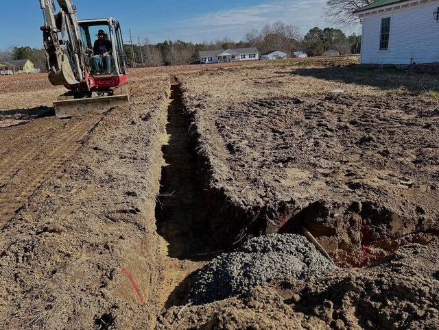 Excavator digging a long trench beside a house on a dirt lot.