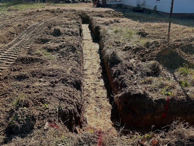 Narrow trench cut through dry dirt beside a grassy field and a building.