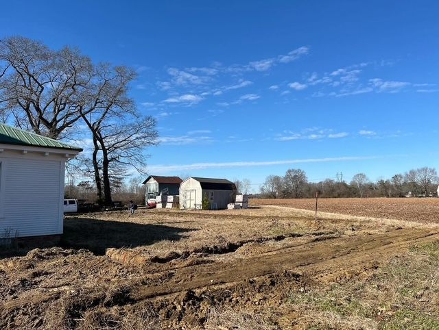 Rural farmyard with a white building, leafless trees, barns, and muddy ground under a blue sky.