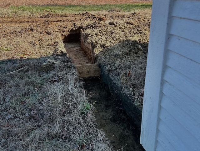 Freshly dug trench in a grassy yard beside a house with a white wall