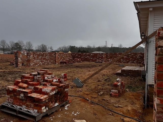 Brick pallets and construction debris on a muddy site beside an unfinished brick building under a gray sky