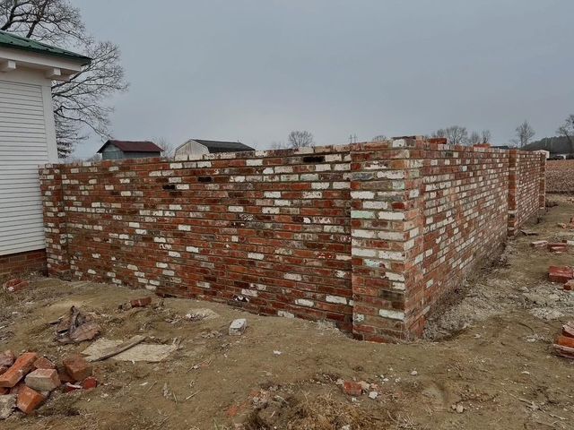 Partially built brick wall structure at a construction site next to a white house, with rubble on the ground