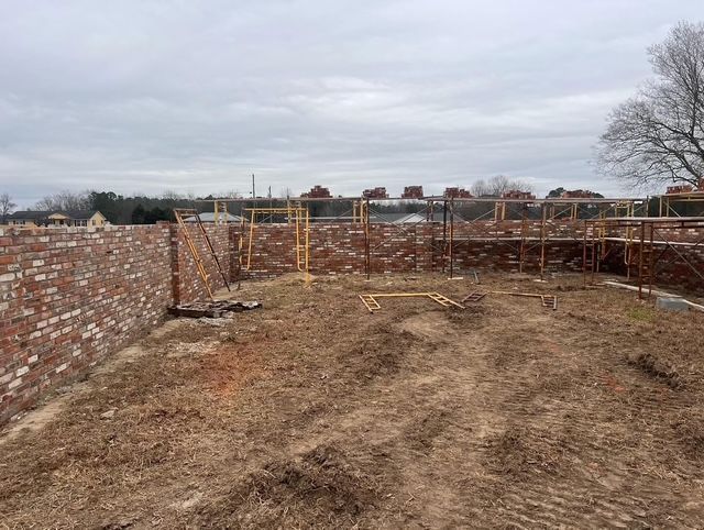 Brick house foundation under construction with wooden scaffolding and a dirt foreground under cloudy skies