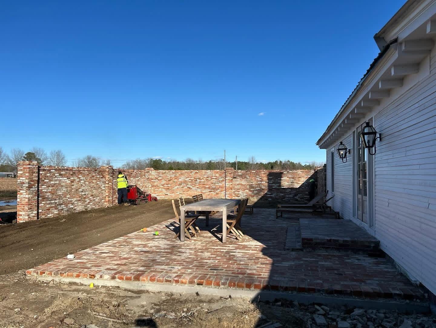 Brick patio and house exterior under construction, with a worker in a neon jacket near a stone wall.