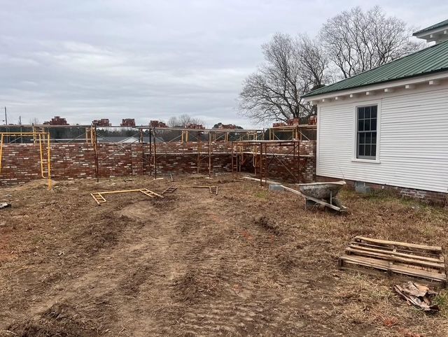 Dirt construction yard beside a white house with green roof, wooden fence, and bare trees under cloudy sky
