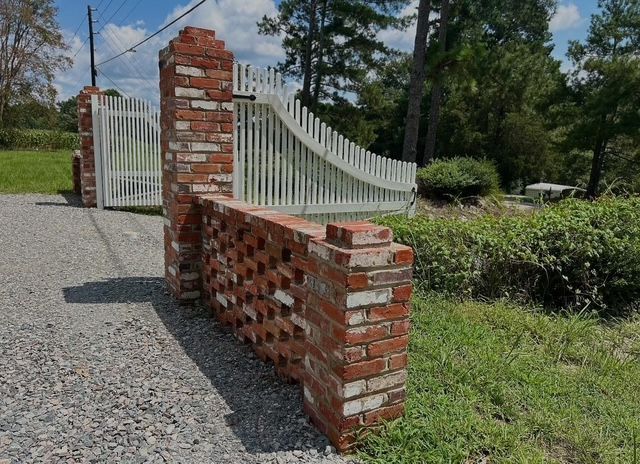 Brick-and-iron gate entrance along a gravel driveway with trees and shrubs nearby