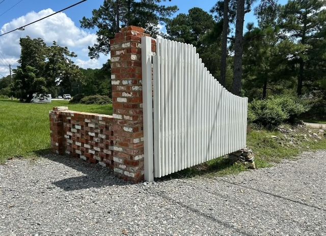 White corrugated metal gate beside a red brick wall on a gravel drive with trees and grass around it