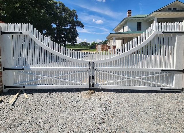 White metal double driveway gate with curved top, closed over a gravel driveway in front of a house