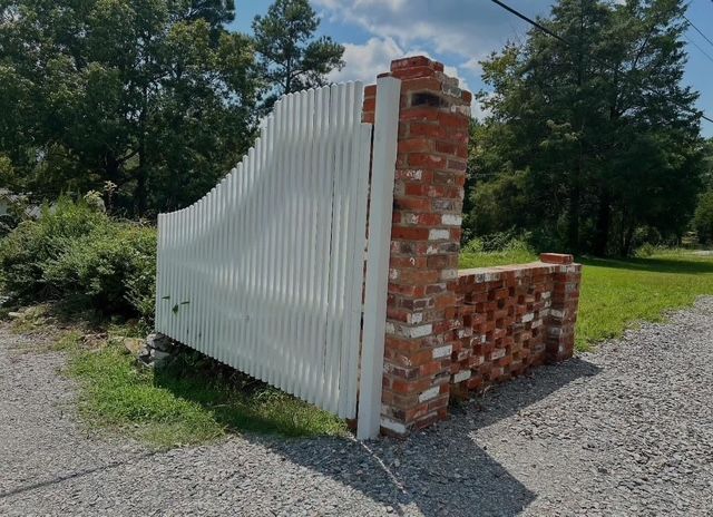 Damaged white fence with a brick end post on a gravel driveway beside grass and trees