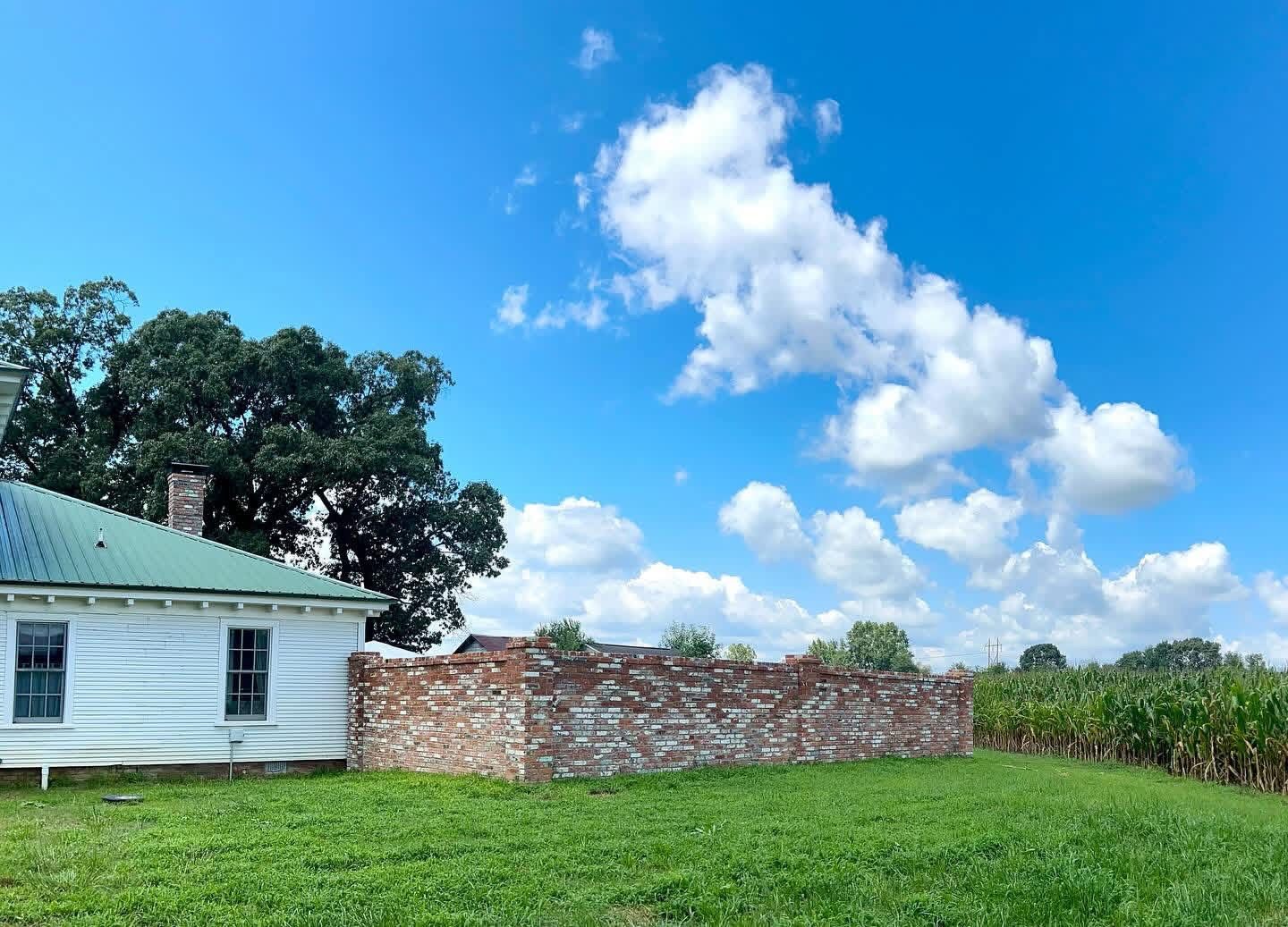 White house with green roof beside a brick wall and cornfield under a bright blue sky with clouds