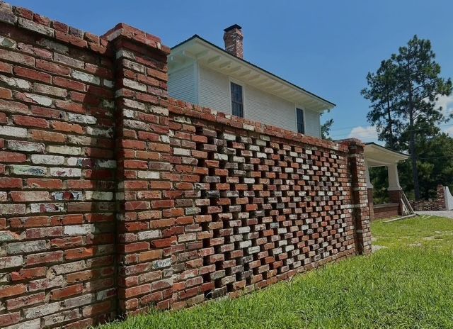 Brick wall with patterned openings in a grassy yard beside a house under a blue sky
