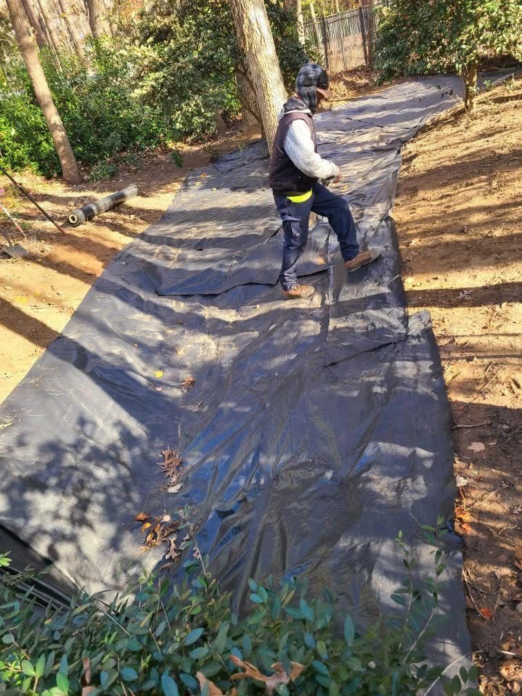 Worker laying black landscape fabric on a sloped garden path in a wooded area