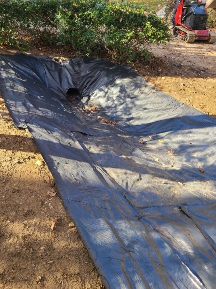 Black tarp-lined ditch or pond with a hole, beside dirt and bushes outdoors