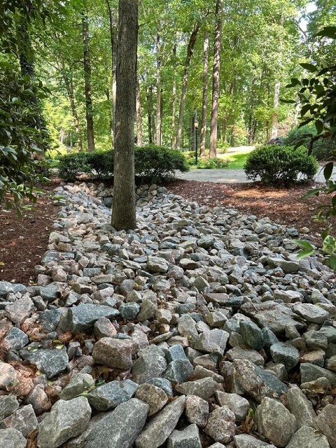 Rock-covered garden bed with a tree, bordered by shrubs and a wooded park path