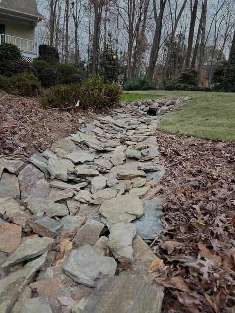 Rock-lined drainage swale beside a lawn and leaf-covered yard near a house