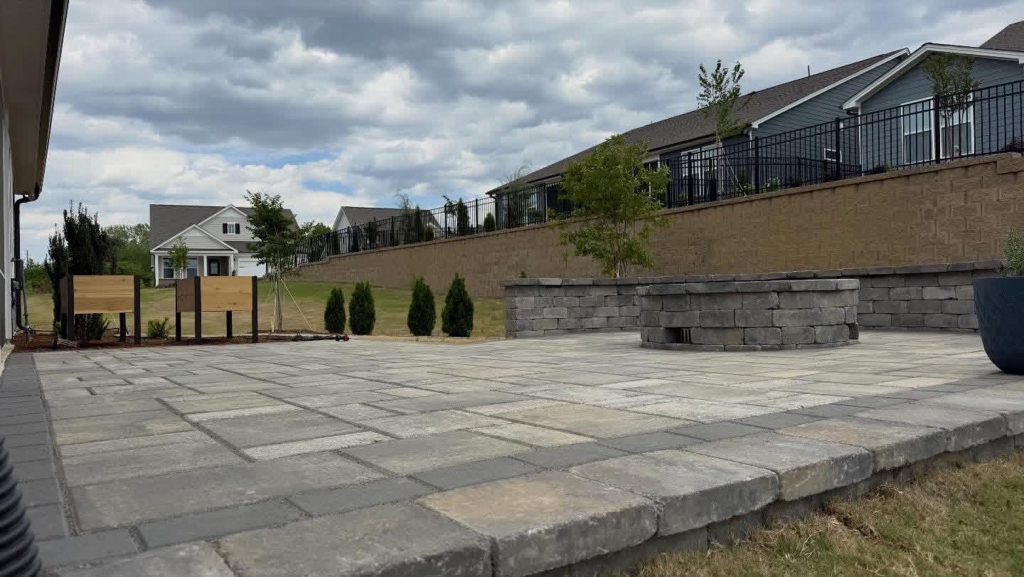 Stone-paved courtyard with retaining wall, small shrubs, and a house with a fenced deck on a cloudy day