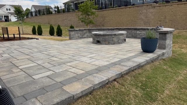 Paver patio with built-in stone fire pit, blue planter, and retaining wall in a landscaped yard.
