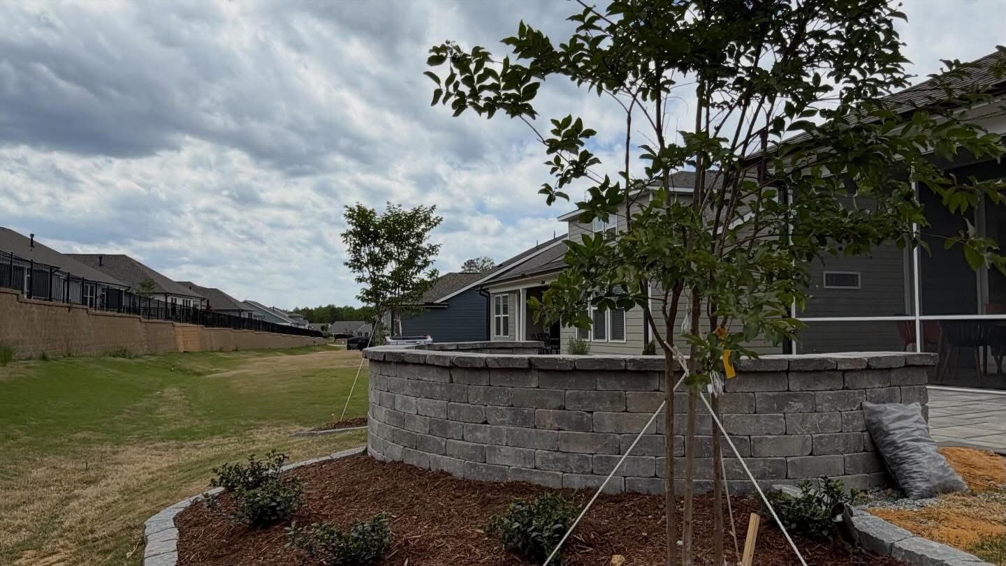 Residential yard with young trees, a stone planter wall, and cloudy sky.