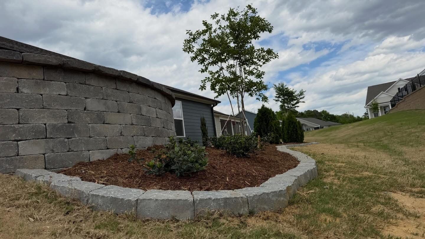 Stone house with a small landscaped garden bed, young tree, and grassy yard under a cloudy sky
