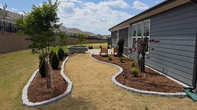 Curved mulched garden beds with young trees beside a gray house and lawn in a suburban backyard