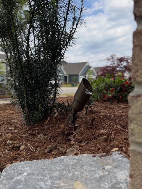 Landscape spotlight in a mulch bed beside shrubs and a house, viewed past a stone edge.