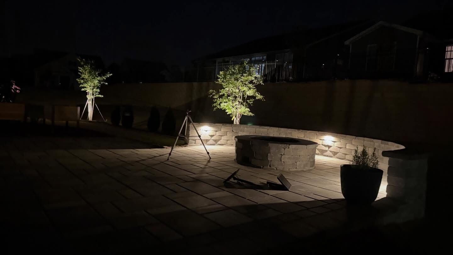 Nighttime patio with string lights, a round stone fire pit, and potted plants against a fence.