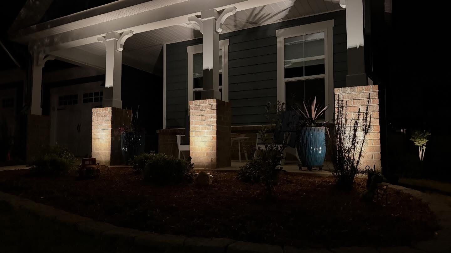 Front porch at night with white columns, lit brick pillars, and potted plants in front of a house