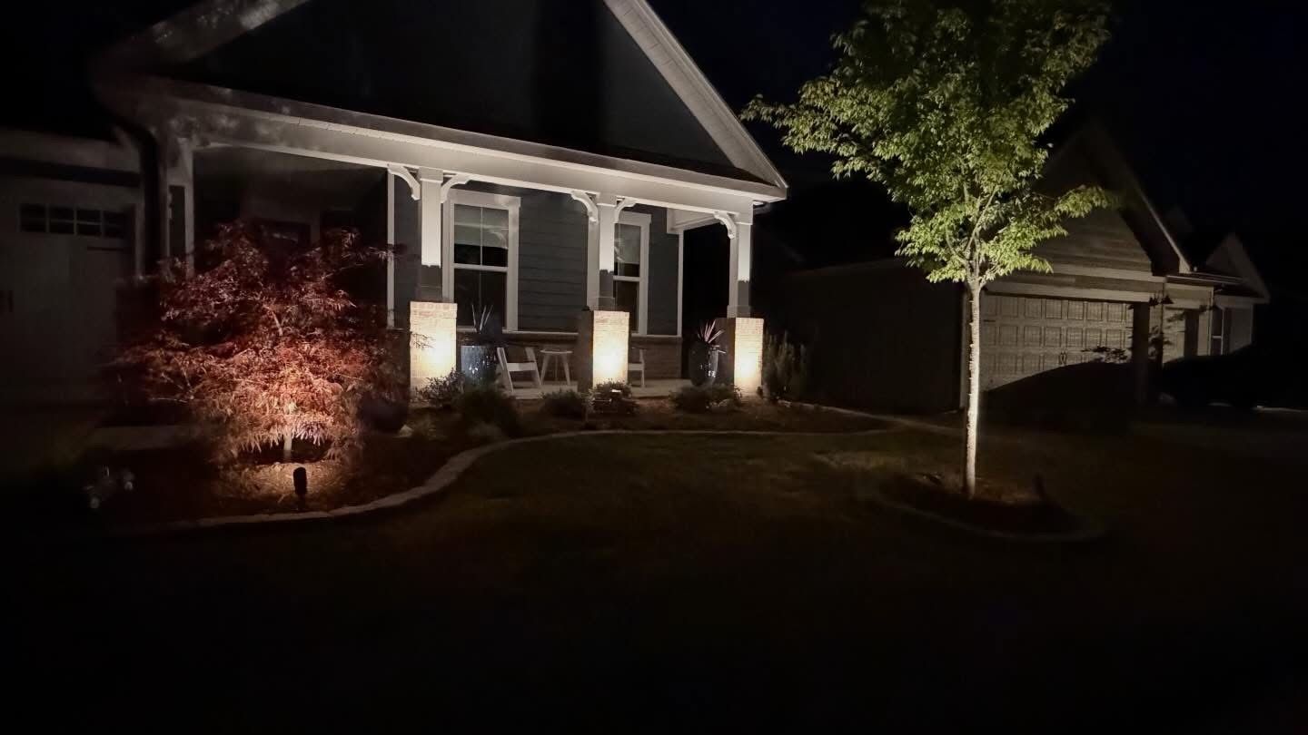Nighttime suburban house with porch lights, a lit tree, and a driveway/garage in front.