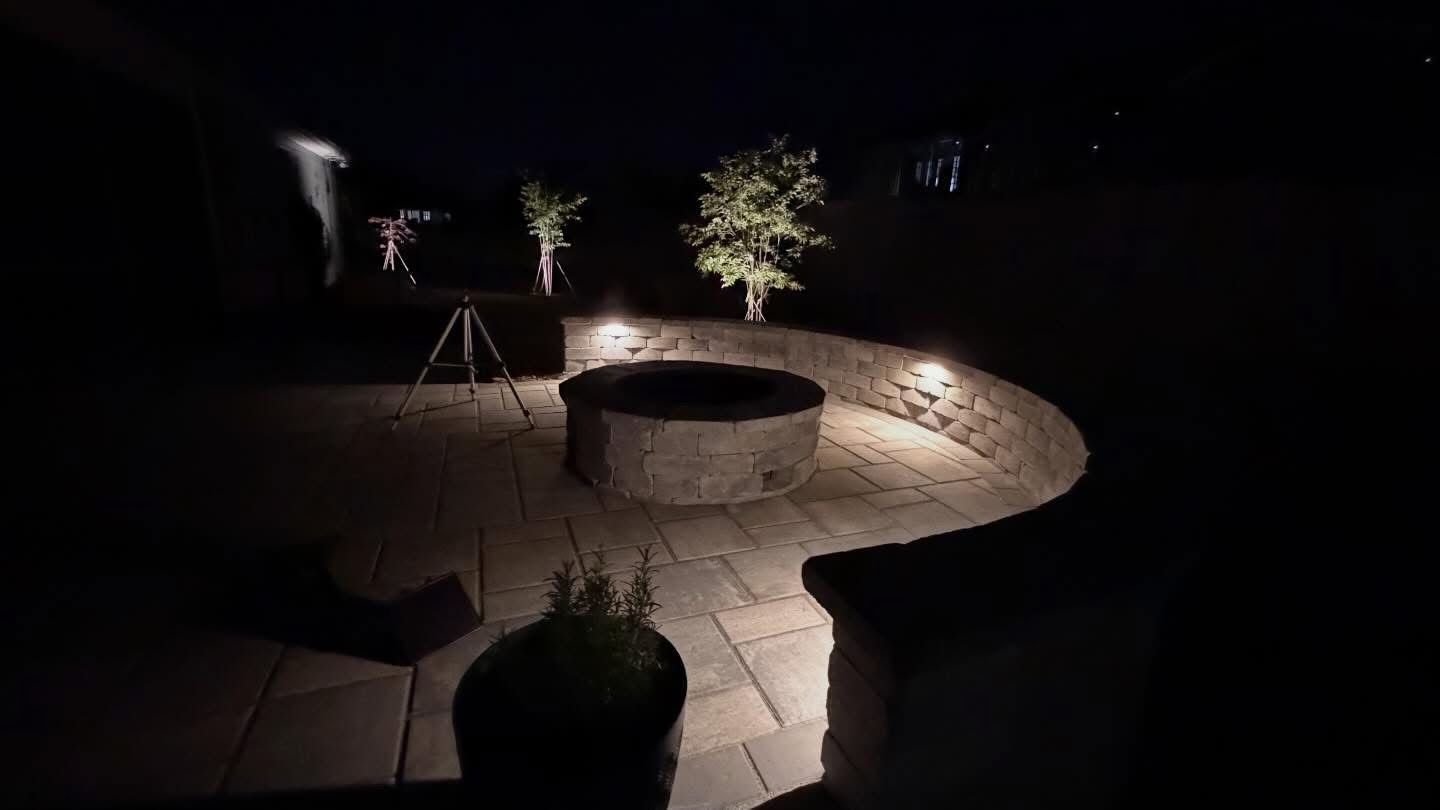Dimly lit stone patio with a round fire pit and potted plants at night
