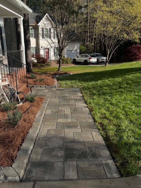 Stone walkway beside a suburban house, bordered by grass and shrubs, leading toward a driveway and parked car.