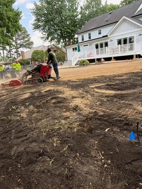 Workers using a red tiller in a dirt yard beside a white house and porch