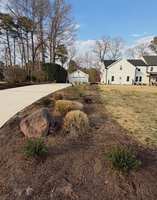 Landscaped garden bed beside a driveway, with rocks, shrubs, and white houses in the background